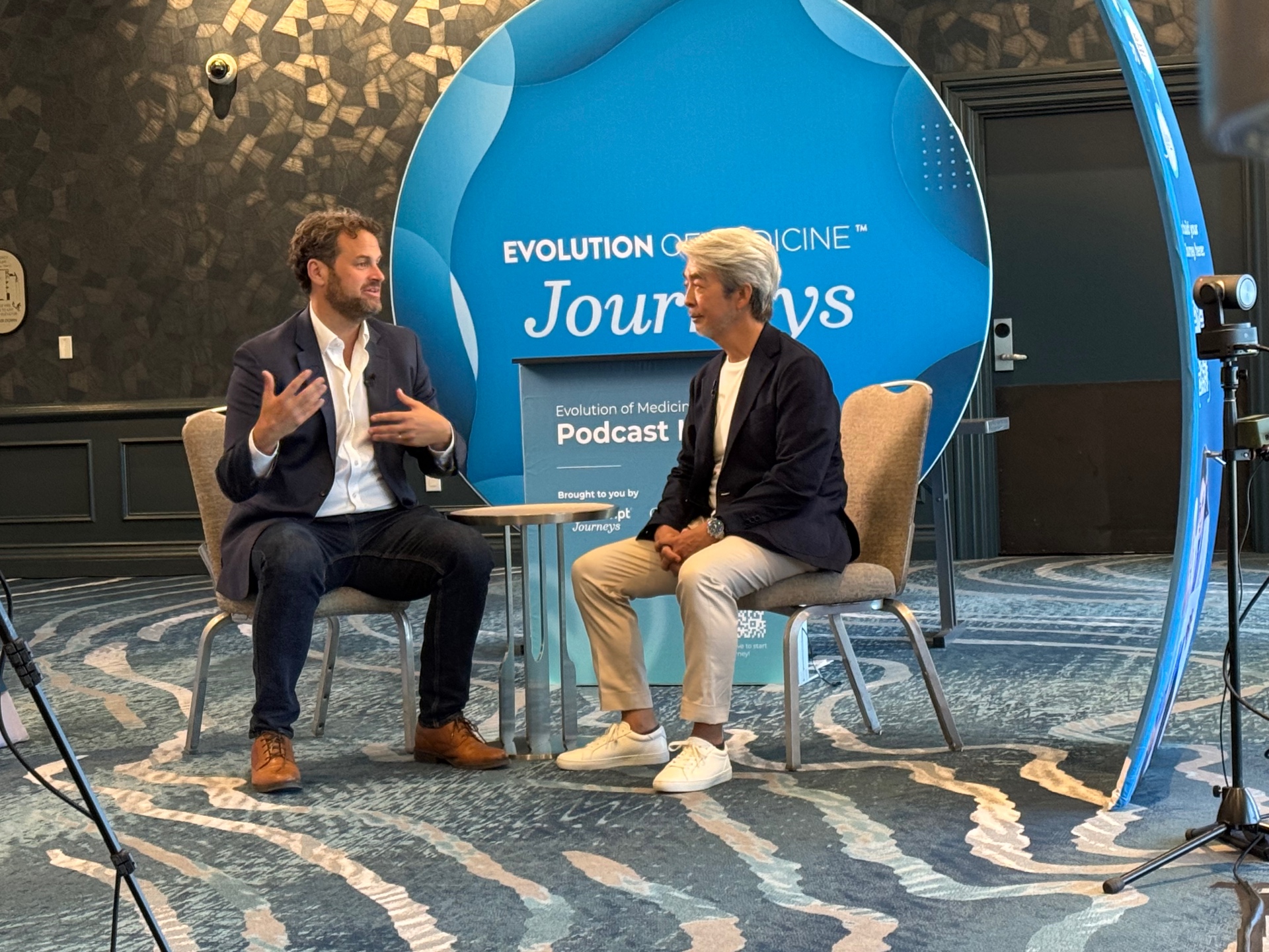 James Maskell and Dr. Kenji Shibuya in conversation in front of the Evolution of Medicine Journeys backdrop